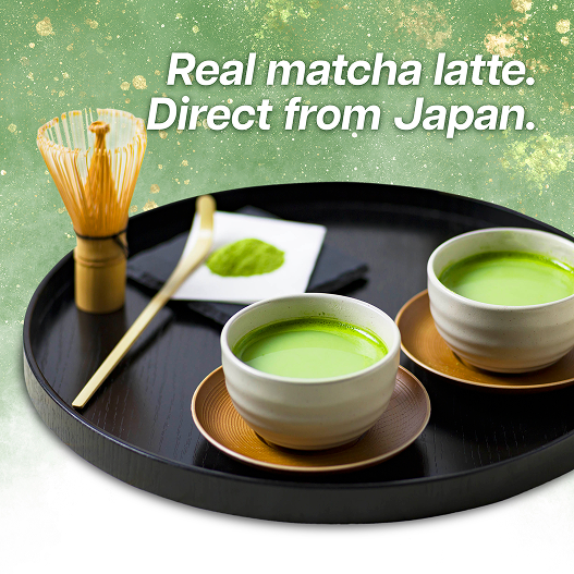 Two cups of matcha latte on a black tray with a whisk and leaf, against a green background.