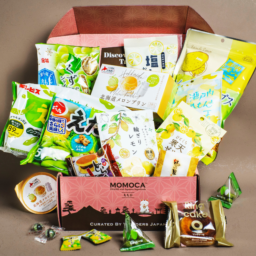 A pink box labeled "Day of the Sea Japanese Snack Box" from MOMOCA is filled with various packaged Japanese snacks, including sweets, cookies, and chips. The snacks have colorful packaging, predominantly in shades of green, yellow, and white.