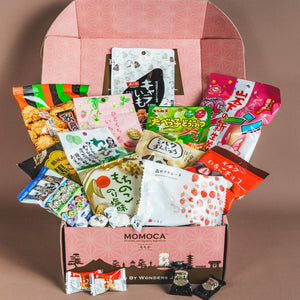 An open Dream Japanese Snack Box by MOMOCA, featuring a pink patterned lid, showcases an assortment of colorful Japanese snacks.