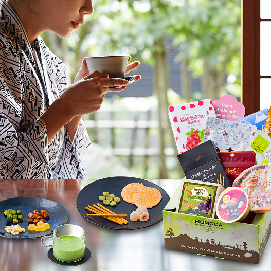 Person drinking from a cup with a table full of snacks and a Momoca box in the foreground.