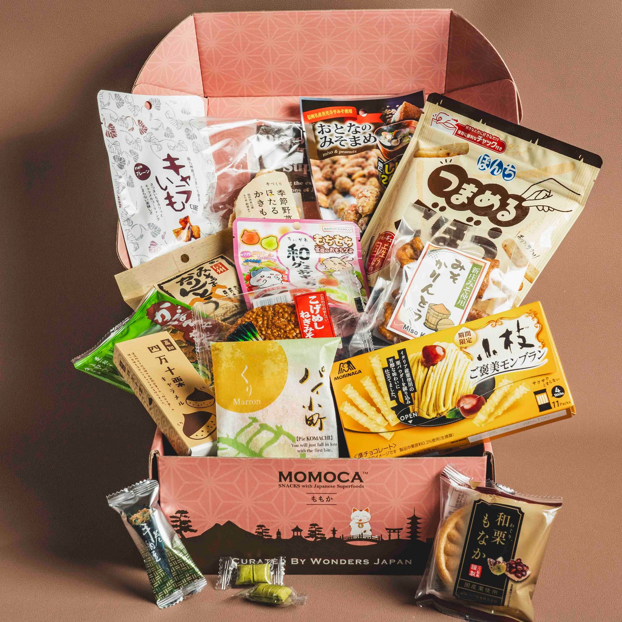 An assortment of Japanese snacks is presented inside an open "Harmony" Japanese Snack Box from MOMOCA, featuring chips, crackers, and sweets. The box lid showcases pink patterns along with the text "Curated by Wonders Japan.