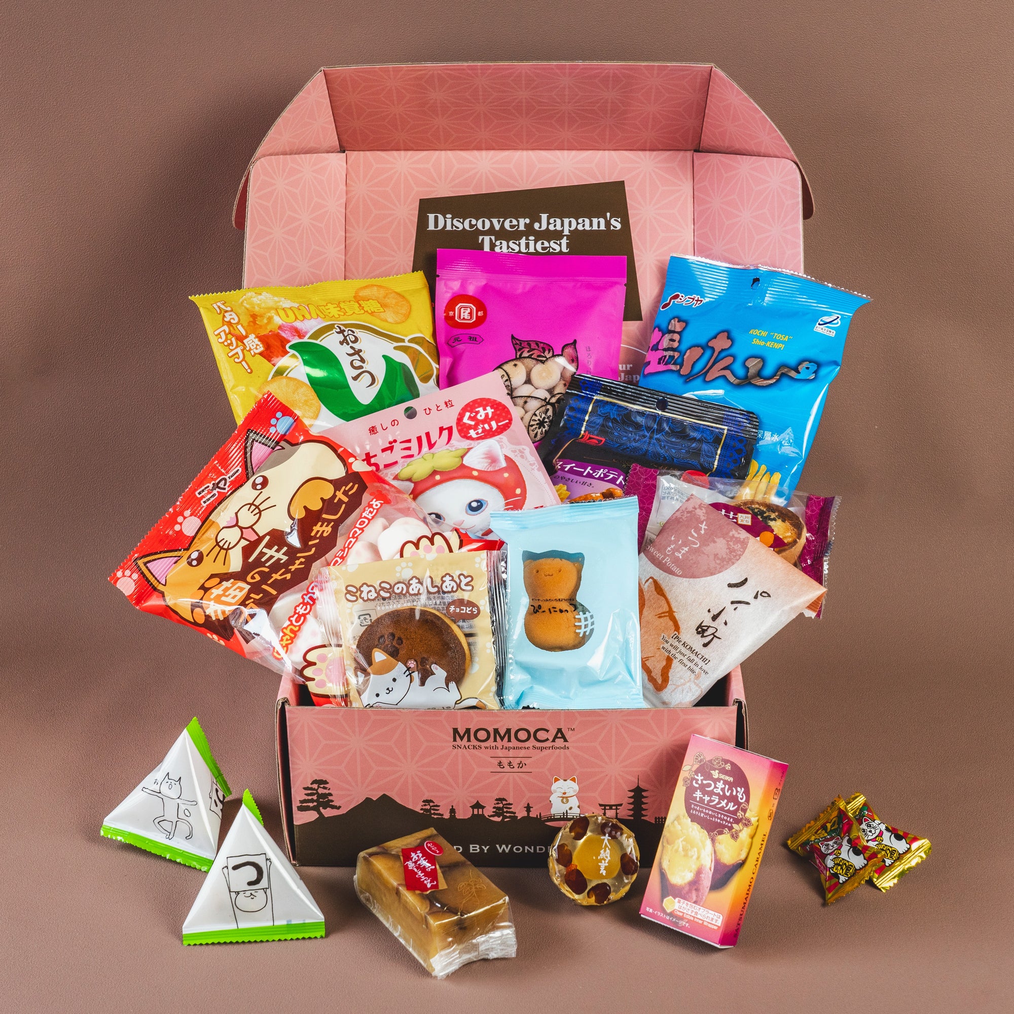 A pink box labeled "Lucky Cat" by MOMOCA is open, revealing a variety of colorful Japanese snacks, including chips, candies, and packaged treats, displayed cohesively inside.