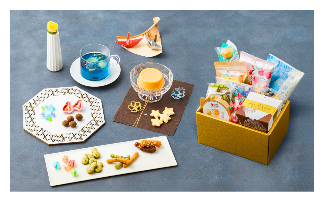 Assorted snacks and treats on a gray surface with decorative plates and a yellow box.