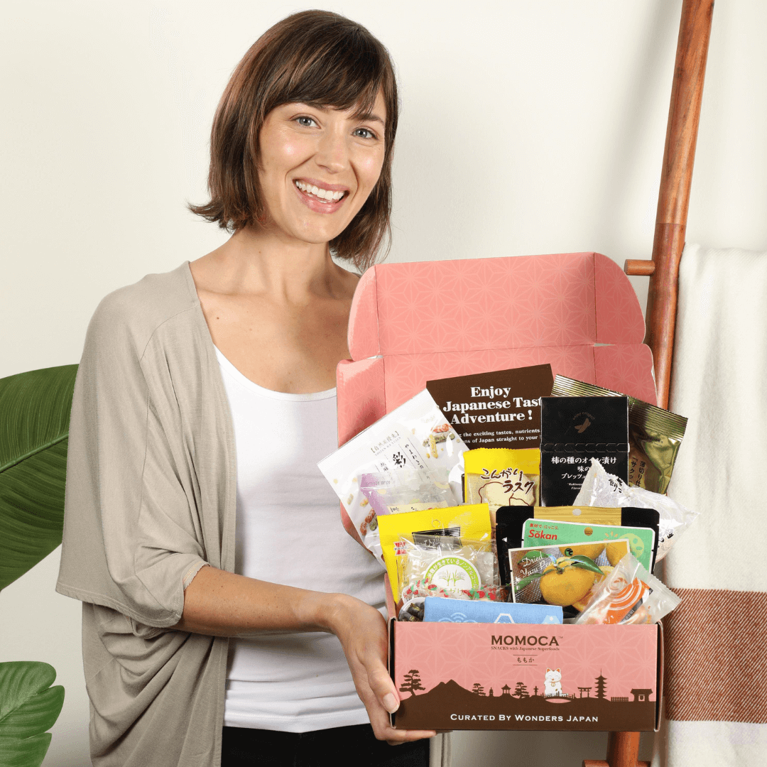 A woman is holding an open pink Wellness Welcome Subscription Box filled with various authentic Japanese snacks and items. The text on the box reads "MOMOCA - Curated by Wonders Japan." The backdrop includes a plant and a towel on a ladder.