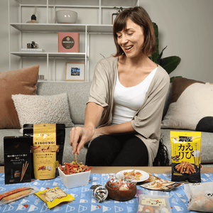 Woman sitting on a couch, smiling while choosing Japanese snacks from various bowls, with Momoca Wellness Welcome Snack Box packages on her side.