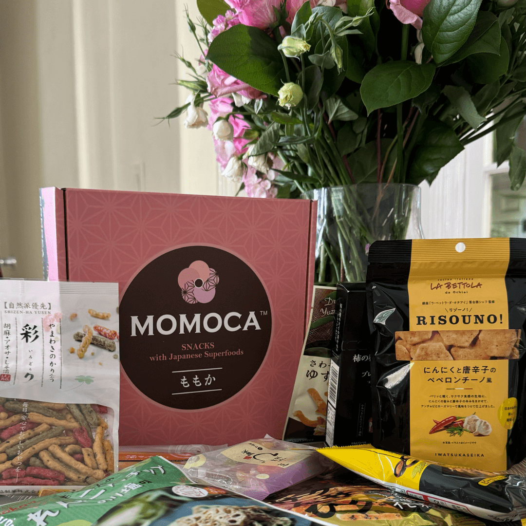 A variety of authentic Japanese snacks from the brand Momoca Wellness Welcome Subscription Box are displayed on a table with a bouquet of pink flowers in the background.