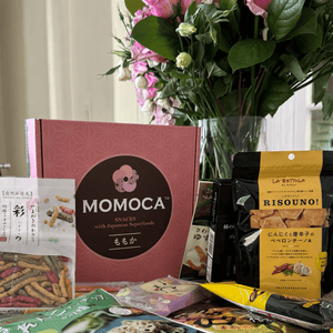 A variety of curated Japanese snacks displayed on a table with a floral bouquet in the background and a Momoca Wellness Welcome Snack Box.