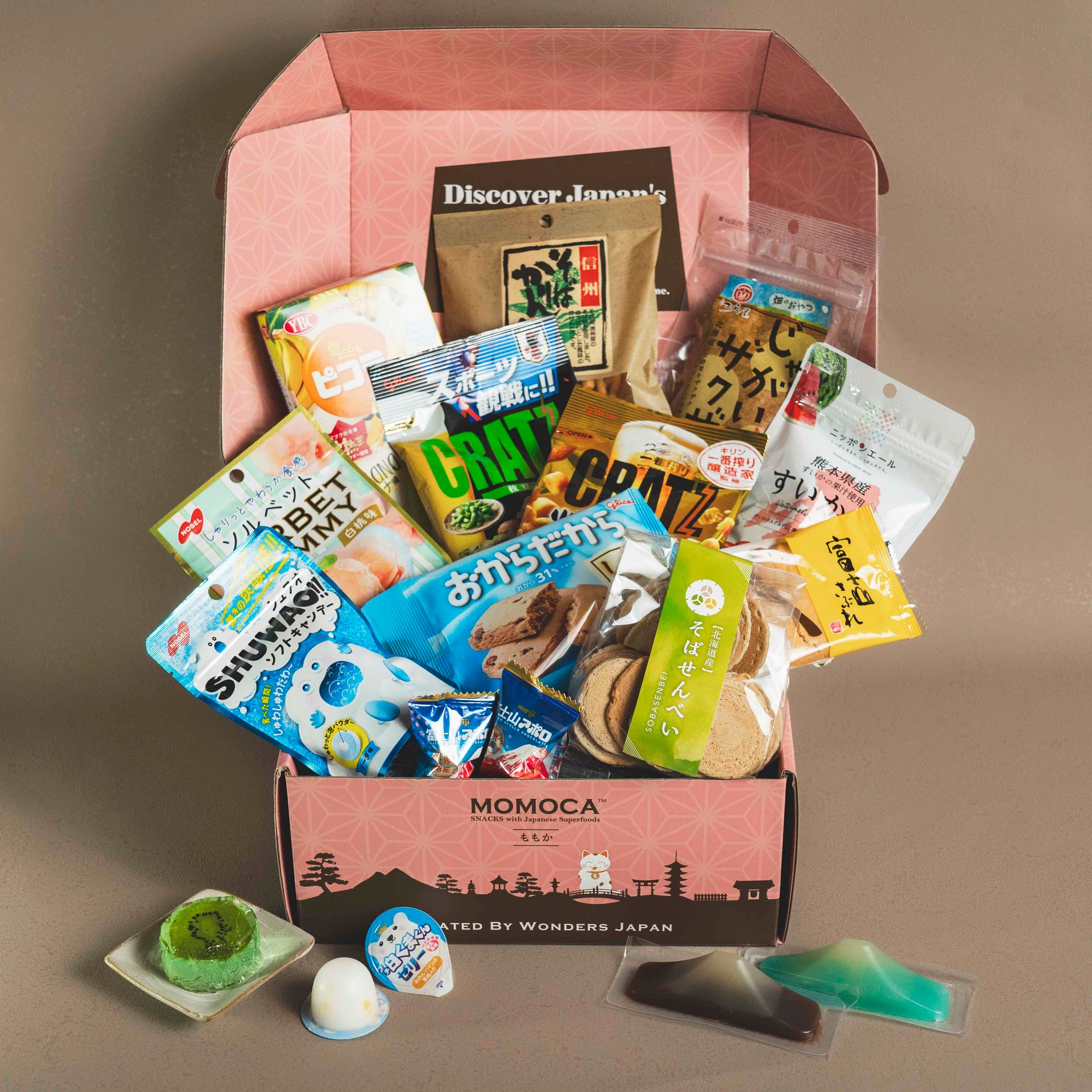 A bright pink box labeled "Mt. Fuji Japanese Snack Box" from MOMOCA is open, revealing an assortment of Japanese snacks, including chips, candies, and cookies, all in various colorful packaging.