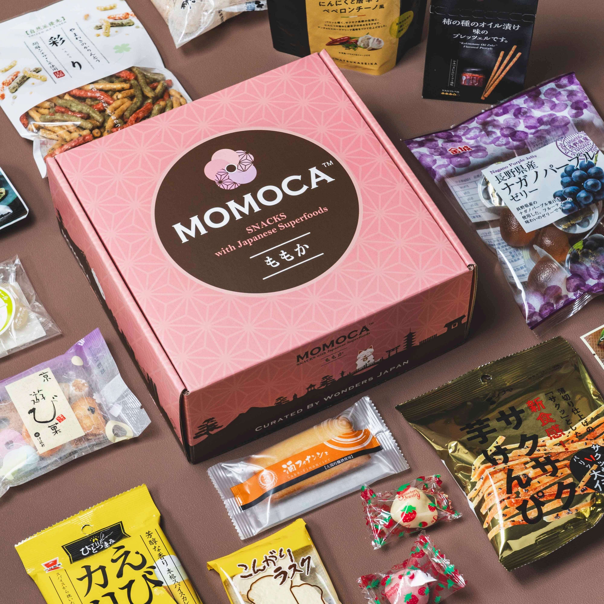 A pink Momoca Wellness Welcome Subscription Box filled with various authentic Japanese snacks and candies is open, displaying its contents on a brown background. A small black dish with a round treat is placed beside it.