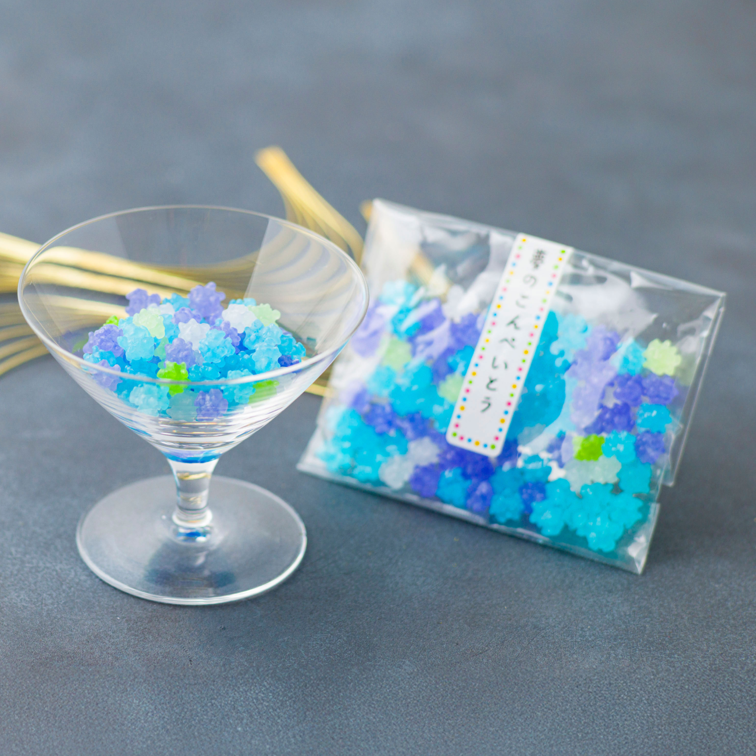 Clear glass with blue and green decorative beads next to a clear bag filled with similar beads on a gray surface.