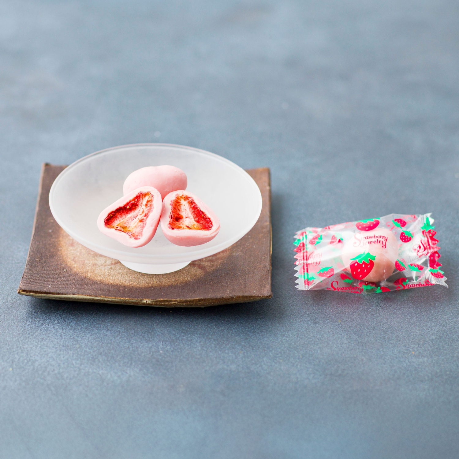 Two pink treats with red centers on a white plate, next to a wrapped treat with strawberry design on a gray background.