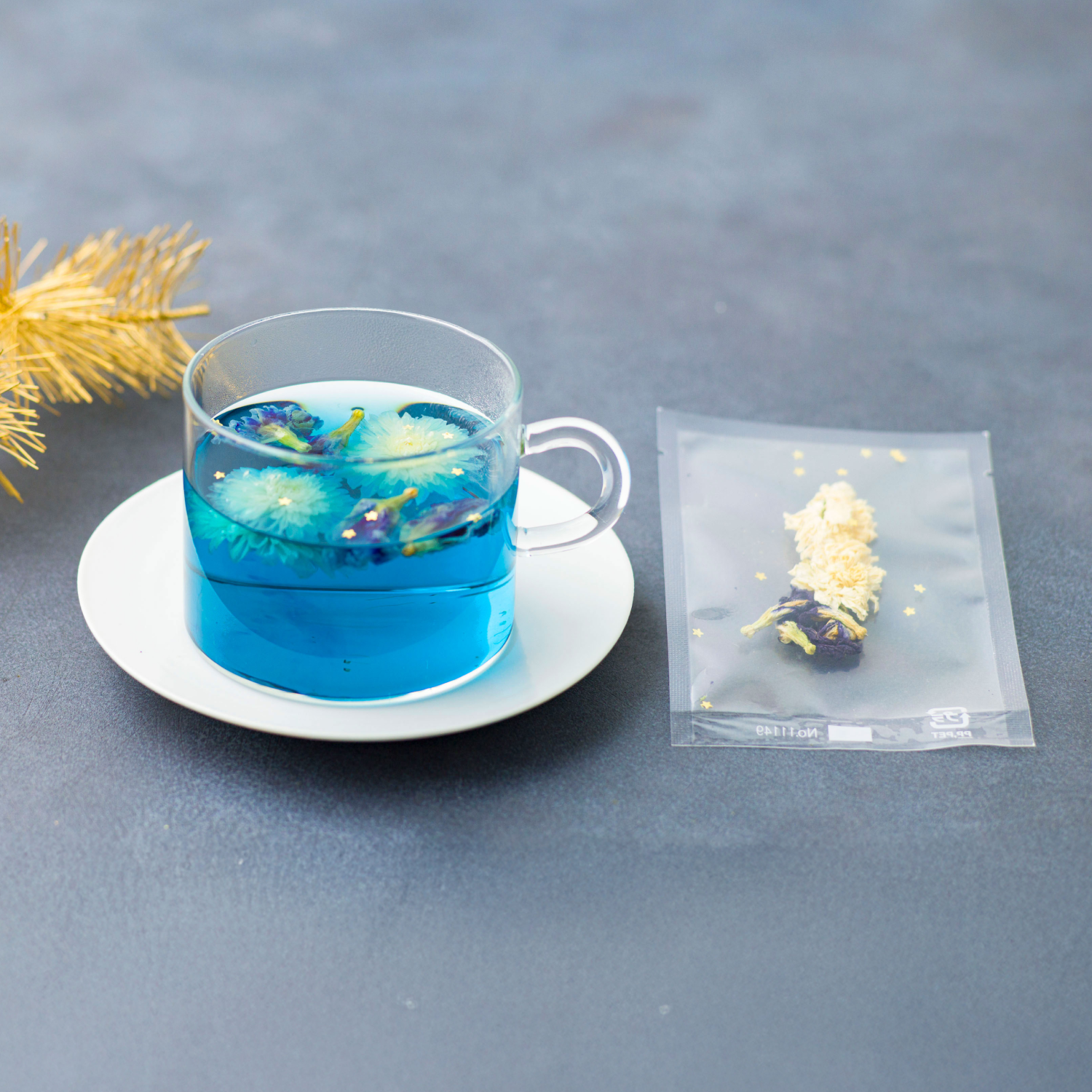 Clear glass mug with blue liquid and floral elements on a white saucer, next to a clear tea bag with yellow flowers on a gray surface.